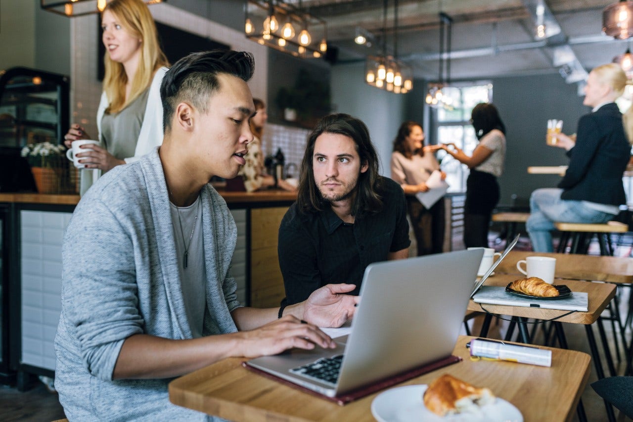 683807932 Two young entrepreneurs sitting in a busy business cafe, working and discussing projects and ideas.