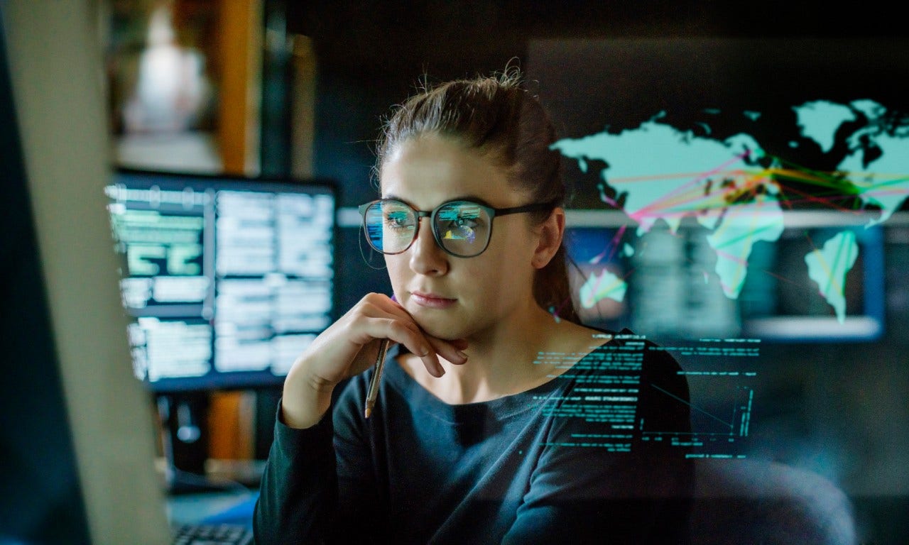 1299152059 Stock image of a young woman, wearing glasses, surrounded by computer monitors in a dark office. In front of her there is a see-through displaying showing a map of the world with some data.