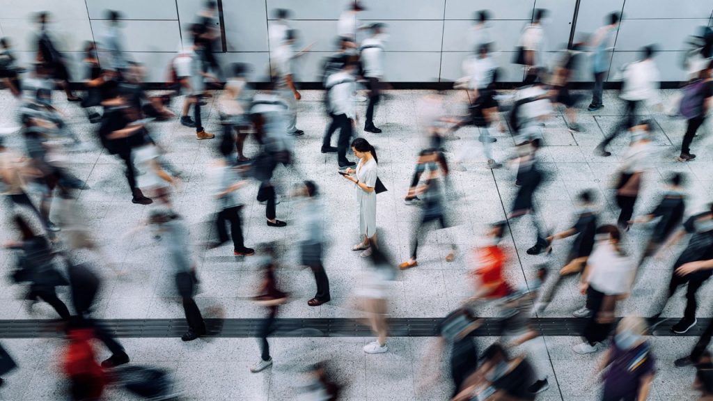1226216285 Young Asian woman using smartphone surrounded by commuters rushing by in subway station during office peak hours in the city