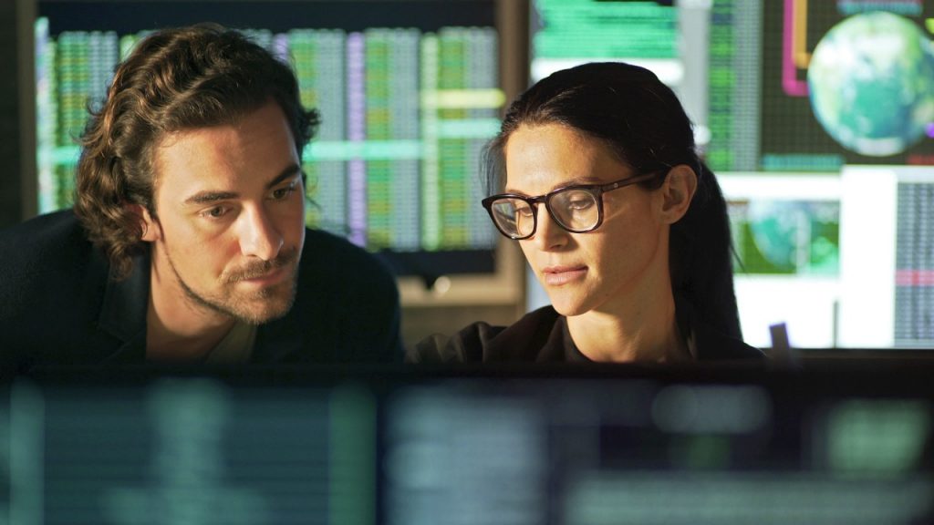 1165363975 Stock photo showing a young man being tutored by a more mature woman. Theyâ re both surrounded by large computer displays showing moving numerical, business & global data.