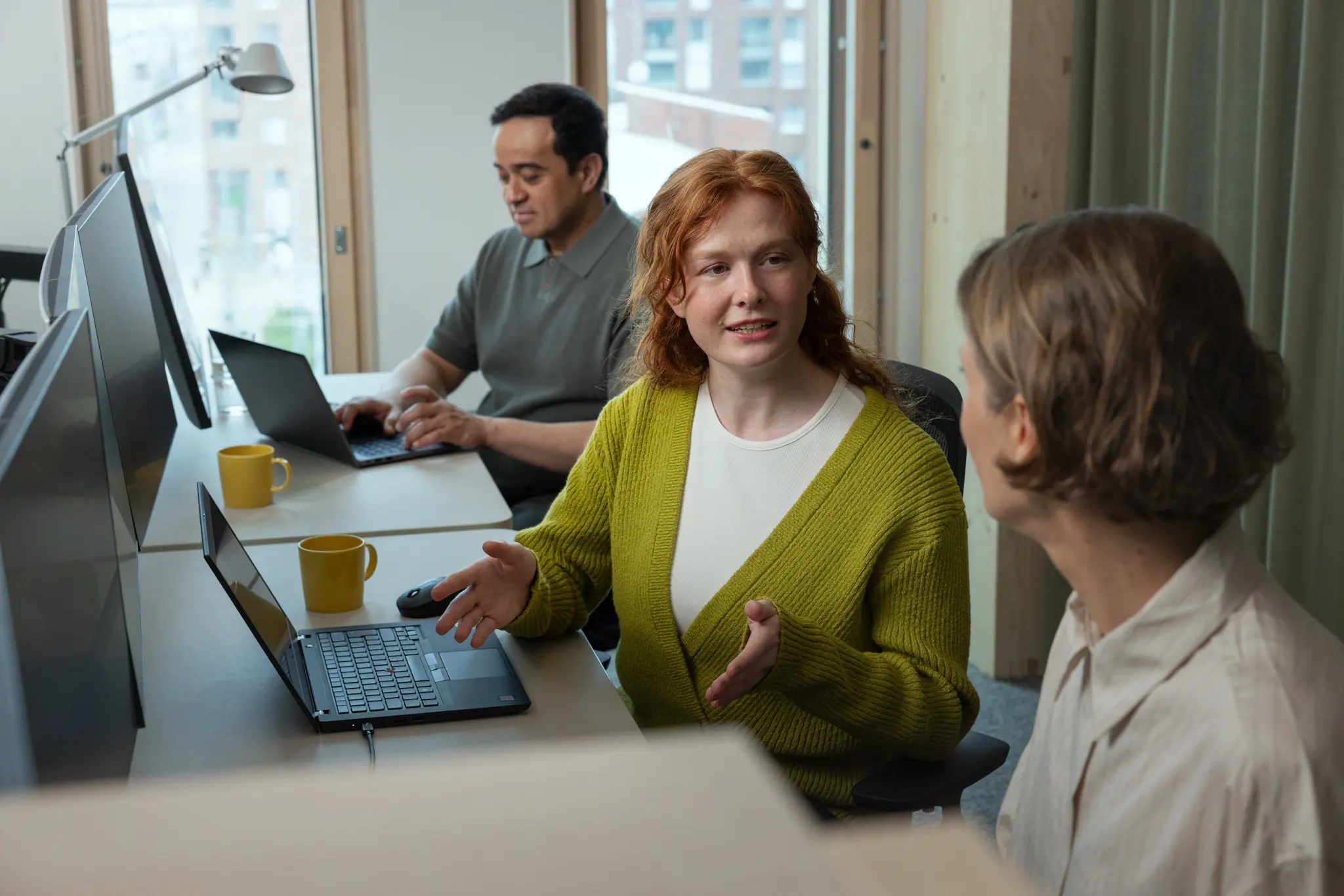 A woman in a green cardigan gestures while talking with a colleague in an open-plan office, a coworker working at dual monitors in the background.