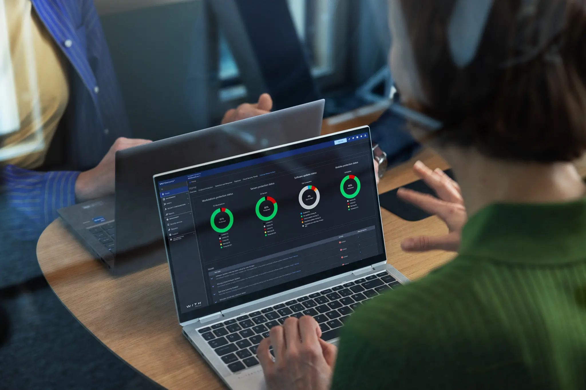 A person in green types on a laptop showing the WithSecure Elements dashboard with colored ring status indicators, a colleague seated across the table.