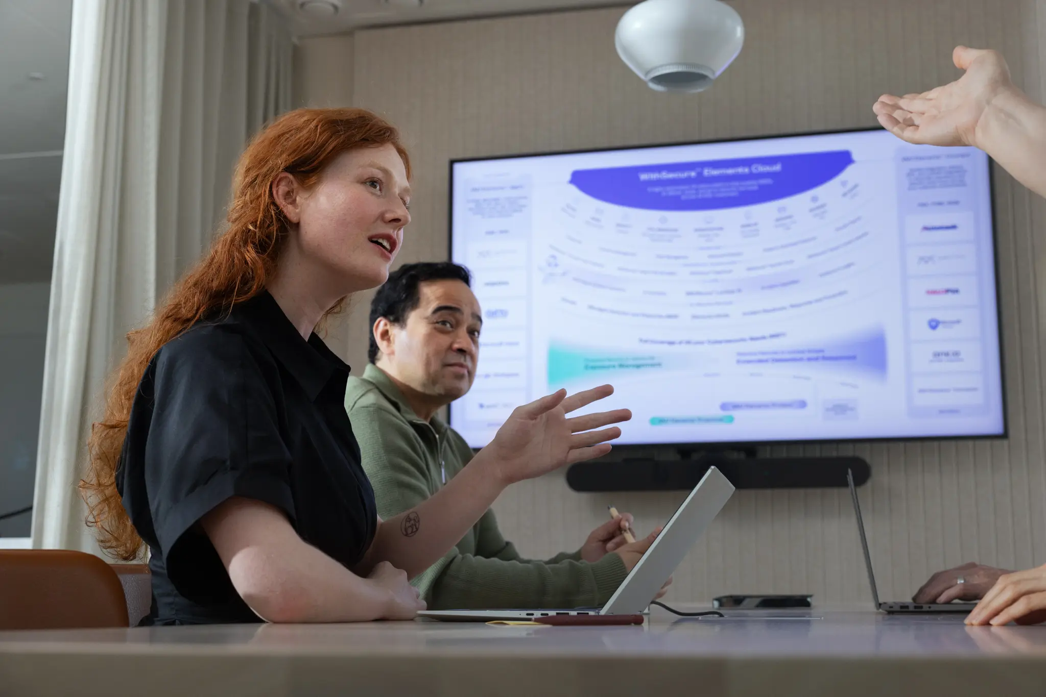 A woman with long red hair gestures while presenting beside a seated colleague, in front of a wall screen showing the WithSecure Elements Cloud architecture diagram.