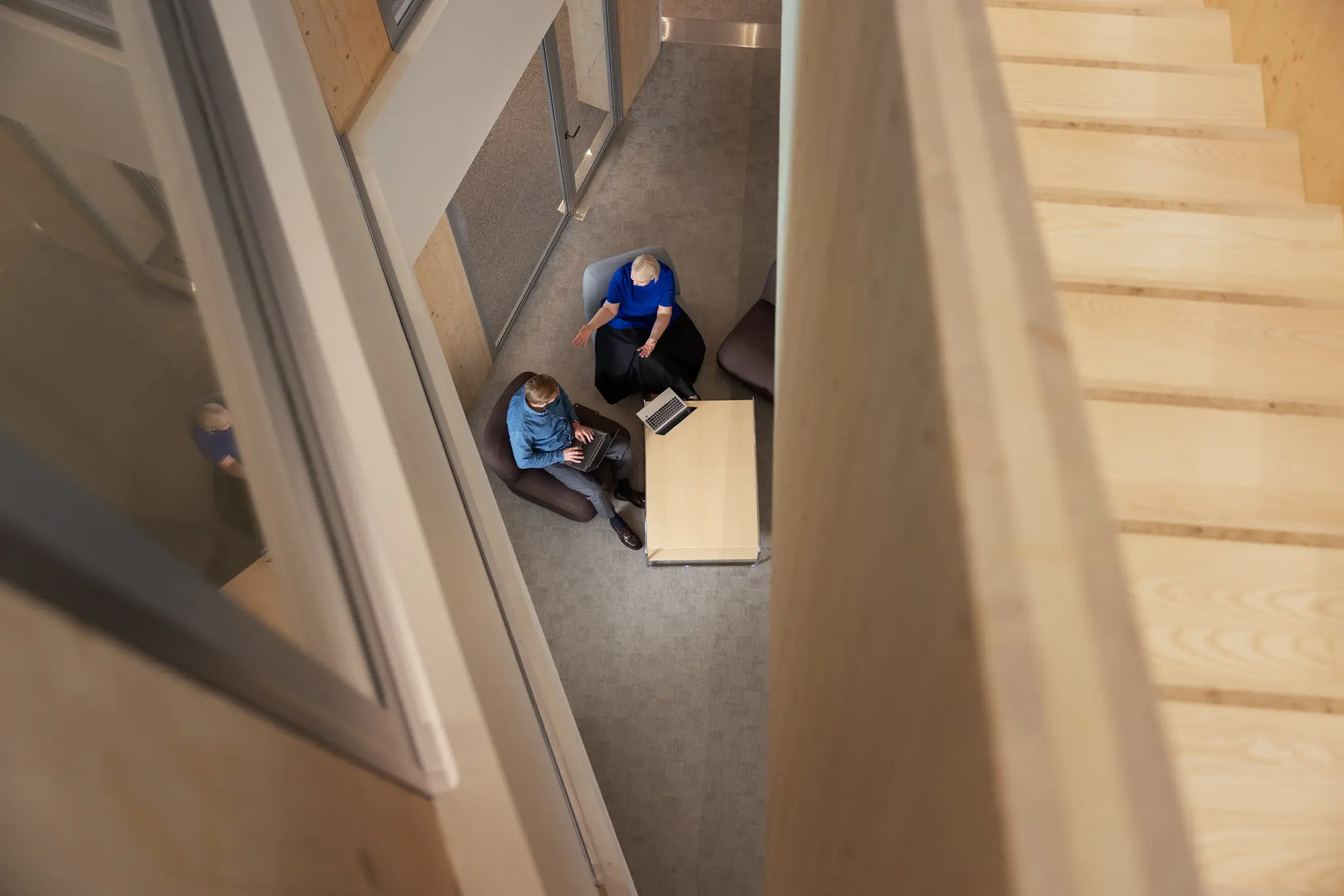 Overhead view down a wooden staircase of two people working on laptops in a casual seating nook below.